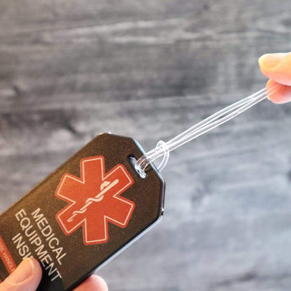 A hand pulls a clear, elastic loop through a hole in a black tag labeled "Medical Equipment Inside," featuring a red star of life symbol. This 2 Pack - Heavy Duty Medical Equipment Luggage Tags CPAP / BIPAP with Plastic Loops ensures your CPAP/BIPAP accessories are easily identifiable during travel.