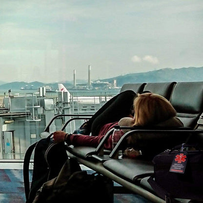 A person is lying down across several chairs in an airport waiting area, using a backpack and neck pillow for comfort. An airplane is visible outside the window, with an industrial landscape beyond it. A 2 Pack - Heavy Duty Medical Equipment Luggage Tags CPAP / BIPAP with Plastic Loops dangles from their bag, ensuring their safety while they rest.