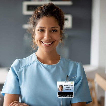 A woman smiles while wearing blue scrubs and a laminated teslin "Pharmacist" Horizontal Badge Buddy with Black Border that reads "Big City Hospital - Cindy Valencia - Pharmacist." She is standing in a professional setting.