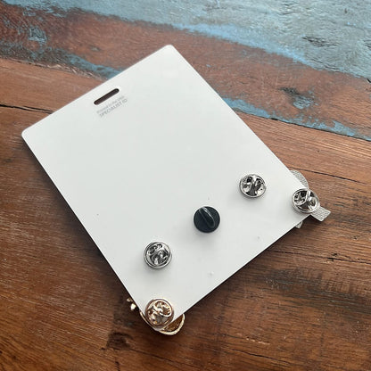 A white rectangular object with four pin-back attachments, including a black and silver screw-like piece, is displayed on a wooden surface. This setup resembles a Pin Buddy Badge for Displaying Pins with Badge Buddies - Horizontal, perfect for organizing your collection.