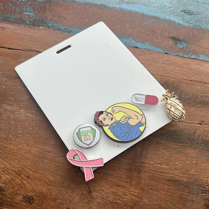 A white Pin Buddy Badge for Displaying Pins with Badge Buddies - Horizontal displays five pins: a pink ribbon, a green USPS logo, a Rosie the Riveter figure, a "She/Her" label, and a gold pineapple. The Pin Buddy Badge for Displaying Pins with Badge Buddies - Horizontal is laid out on a wooden surface.