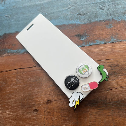 A white rectangular Pin Buddy for Displaying Pins with Badge Buddies - Vertical serves as a pin holder with three pin buttons and two dangling pins attached, resting on a wooden surface. The enamel pins include a shield, a pill, a dinosaur, a lightning bolt, and a cloud.