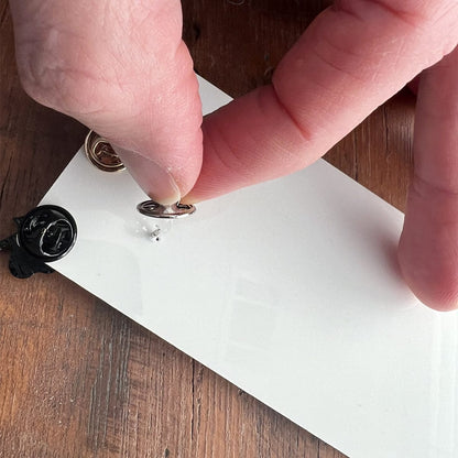 A close-up of a hand holding a loop of wire against a white panel on a wooden surface, with small hardware pieces and Pin Buddy for Displaying Pins with Badge Buddies - Vertical nearby.