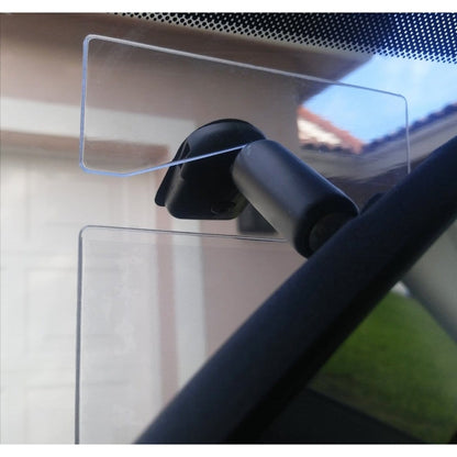 A Specialist ID Handicap Hang Tag Holder (SPID-1120), a transparent rectangular mount doubling as a clear vinyl handicap holder, is attached to a car windshield, secured with a black suction cup and adjustable arm. A blurred background shows a garage and house.