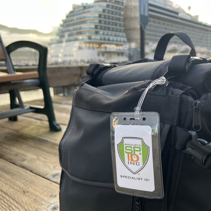 A close-up of a black backpack with a Clear Plastic Luggage Identification Tags with Loops Included - Business Card or Photo Insert (Easy Access) tag attached, set on a wooden pier with a large cruise ship in the background, highlights its secure worm loops for additional safety.
