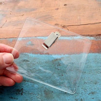 A hand holding a 4 x 3 Horizontal Event Badge Holder with Clothing Friendly Bulldog Clip - Clear Vinyl, Horizontal & Durable (SPID-1440) against a wooden surface with blue paint.