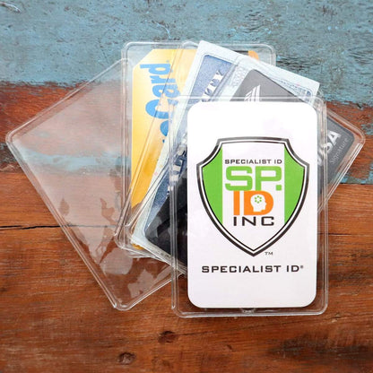 A Clear Vinyl Business Card Holder - Medicare Card Protective Sleeve (1840-3505) containing several identification cards is placed on a wooden surface with a blue backdrop. The visible card features the logo and name "Specialist ID, Inc.