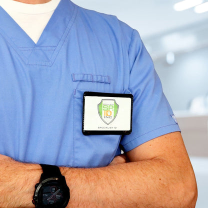 Close-up of a person in a light blue scrub top with a smartwatch on their wrist and a badge holder clipped to the shirt, displaying a badge from Specialist ID Inc. The clear vinyl front of the Horizontal Magnetic Badge Holder (P/N 1835-1050) ensures easy visibility, while strong magnets keep it securely in place. Arms are crossed.
