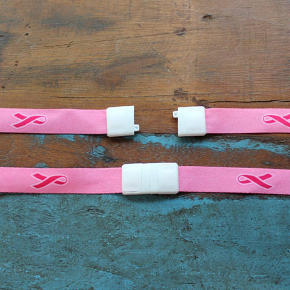 Two pink straps with pink ribbon symbols and white plastic clasps, perfect as a Pink Ribbon Awareness Wide Flat Breakaway Lanyard w/ Swivel Hook 2138-5286, are laid out on a wooden and blue surface.