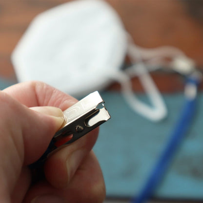 Close-up of a hand holding a silver nail clipper with a blurred white face mask, a blue object, and a 5 Pack Double Ended Lanyards with Bulldog Clips - Comfortable Premium Economy Neck Strap Badge Holder for Events in the background.