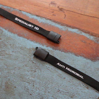 Two black lanyards with white text on a textured surface. One lanyard reads "SPECIALIST ID" and the other reads "ANTIMICROBIAL LANYARD WITH TWO CLIPS - SHORT LENGTH DOUBLE CLIP LANYARD WITH SAFETY BREAKAWAY - 25 INCH PERFECT SIZE FOR STUDENTS & ADULTS (SPID-2360)," showcasing nanoparticle-coated fibers for added germ-inhibiting protection.