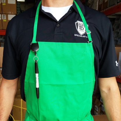 A person wearing a green apron over a black polo shirt with a logo badge, accompanied by a Retractable Pull Badge Reel Pen / Pencil Holder with Carabiner Clip SPID-3280. A pen is secured to the apron with a clip. Boxes and shelves are visible in the background.