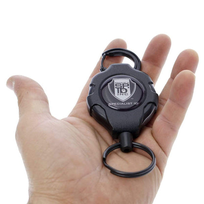 A hand holding a Heavy Duty Retractable Ratchit Keychain Tether Reel for Multiple Keys with Clip and Locking Feature, featuring a logo and the text "Specialist ID Inc" on the device.