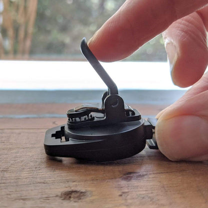 Fingers are adjusting a black folding mechanism on a wooden surface, resembling the precision needed when handling MRI Safe Badge Reel - Non-Ferrous Metal Retractable Badge Clips with No Twist ID Holder Clip for Imaging Room Techs & Nurses. The blurred background adds to the focus on the intricacies of the adjustment.
