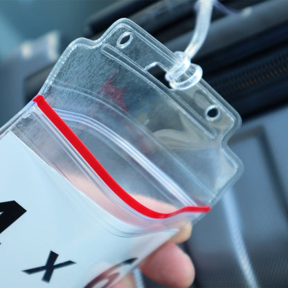 Close-up of a clear plastic ziplock bag with a red seal and a lanyard attached, held by someone's hand in front of a gray suitcase, showcasing its use as the 2 Pack - Extra Large Luggage Tags - Clear 4x6 Heavy Duty Cruise Ship ID Tag / Golf Bag Travel Document Holder with 9" Plastic Loop.