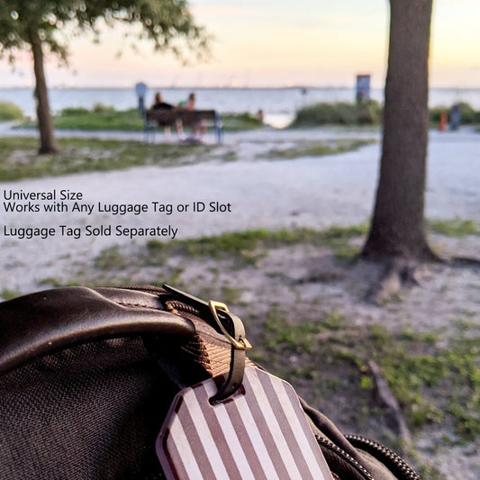 Close-up of a black bag with a striped luggage tag and a Black Genuine Leather Luggage Strap W/ Brass-Plated Buckle, 3 Holes 2420-1041 featuring adjustable holes and a brass-plated buckle, set against an outdoor background with trees and a blurred view of water and sky. Text reads "Universal Size, Works with Any Luggage Tag or ID Slot. Luggage Tag Sold Separately.