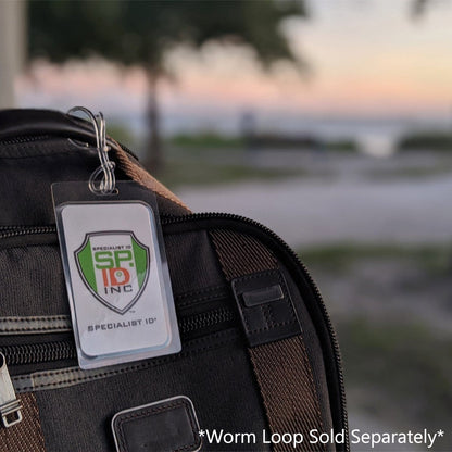 Close-up of a luggage tag attached to a brown suitcase, featuring the logo "Specialist ID SP ID Inc." with a blurred outdoor background at sunset. Text reads: "*7 Mil Medium Duty Laminating Pouch for 2.5" x 4.25" Luggage Tags or ID with Vertical Slot Hole Sold Separately*". Perfect for DIY craft enthusiasts who enjoy personalizing their travel accessories.
