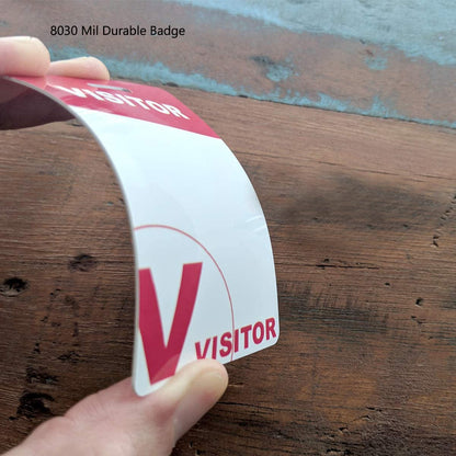 A hand holds a bent visitor badge labeled "VISITOR" on a wooden surface. The badge features a white background with a red "V" and red header. Text on the image says "8030 Mil Durable Badge." These Visitor Badges with Lanyard - Secure Guest ID Passes for Events and Offices ensure secure identification.