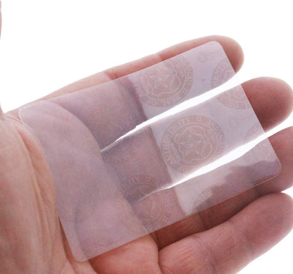 A hand holds a transparent security hologram card with faint circular emblems visible, featuring a Self Adhesive Holographic Overlay - Shield and Key for enhanced anti-counterfeiting protection.
