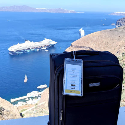 A black suitcase with Premium Vinyl Cruise Ship Luggage Tag Holders with 9" Worm Loops Attach to Your Suitcases, Bag Handles, and Carry On Luggage - 3 1/2 x 7" Clear Protective Insert Sleeves by Specialist ID stands on a balcony overlooking a scenic view of the ocean with two cruise ships near the shore; islands and rugged coastline are visible in the background.