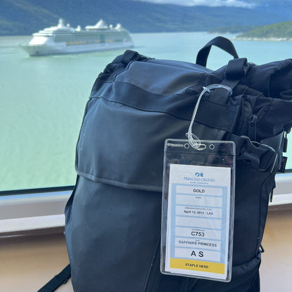 A black backpack with a Premium Vinyl Cruise Ship Luggage Tag Holders with 9" Worm Loops Attach to Your Suitcases, Bag Handles, and Carry On Luggage - 3 1/2 x 7" Clear Protective Insert Sleeves by Specialist ID sits on a windowsill. A large cruise ship sails on a blue-green body of water, framed by a mountainous landscape in the background.