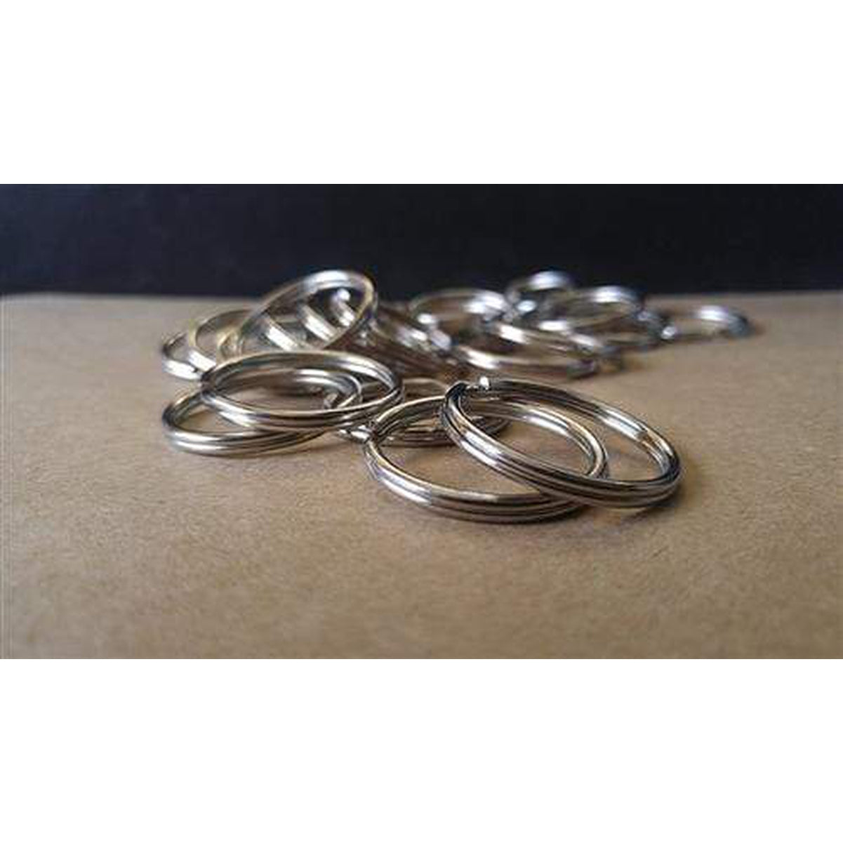 A pile of 25 Premium Extra Large 1 1/4" Split Rings (SPID-9240), crafted from heat treated steel, is scattered on a brown surface against a black background.
