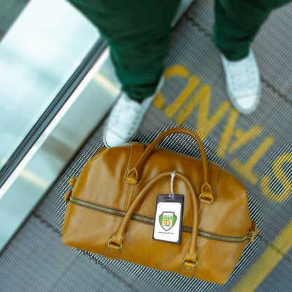 A person in green pants and white shoes stands on an escalator, holding a tan leather handbag with a Color Coded Backpack ID Tags with Loop - Semi Rigid Holds a Business Card or Included Identification Card (1845-200X) attached. The escalator's step has the word "STAND" partially visible.