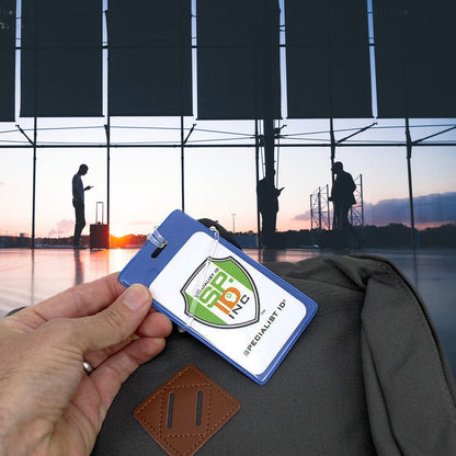 A person holds an ID badge with a "Specialist ID" logo, which doubles as a Color Coded Backpack ID Tags with Loop - Semi Rigid Holds a Business Card or Included Identification Card (1845-200X), in front of a windowed airport scene with people and luggage silhouettes during sunrise or sunset.