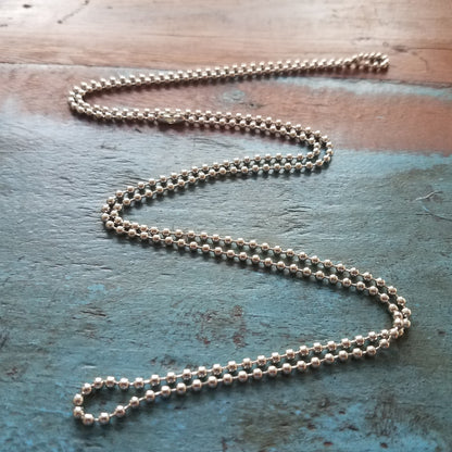 A 2125-1000 Nickel-Plated Steel 24" Beaded Neck Chain is laid out in a wavy pattern on a rustic wooden surface with a weathered blue and brown finish.