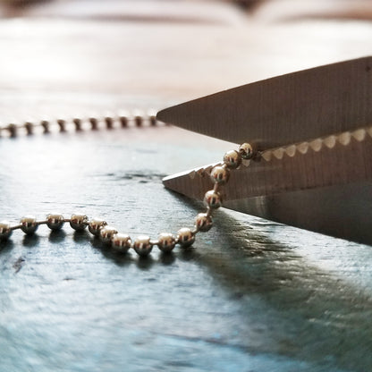 Close-up of scissors cutting a durable accessory, a Nickel-Plated Steel Beaded Neck Chain, Length 30" (762mm) (P/N 2125-1500), placed on a textured surface. Perfect for jewelry craft projects.
