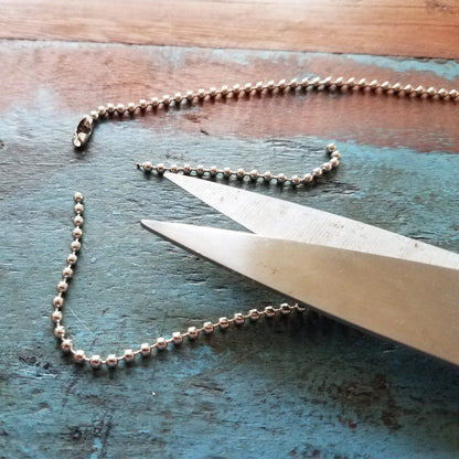 A pair of scissors effortlessly cutting through a Nickel-Plated Steel Beaded Neck Chain, Length 36" (P/N 2125-2000), designed for ID badge applications, on a textured surface.
