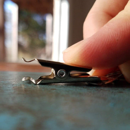 Close-up of a hand holding a metal staple remover on a blue surface with a blurred background, next to Reusable Visitor Passes with Clothing Friendly Badge Clip.