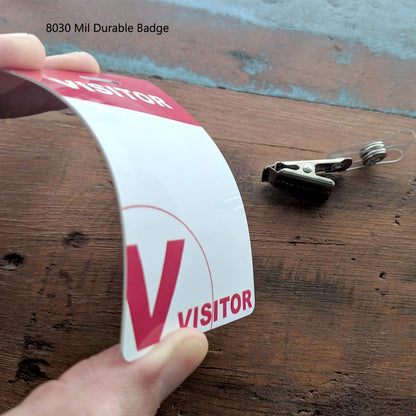 Hand holding a flexible, reusable ID badge with "VISITOR" printed on it next to a metal and plastic clip on a wooden surface. Text on image reads "Reusable Visitor Passes with Clothing Friendly Badge Clip.
