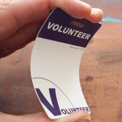 Close-up of a hand holding a bent badge from the 10 Pack - Volunteer Lanyard with Badge Set - Pre-Printed Volunteer Design - Dual Sided, with a purple and white color scheme, attached to a lanyard. The badge clearly displays "VOLUNTEER" in bold letters.