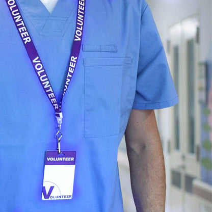 A person wearing a blue scrub shirt with a lanyard and a badge labeled "Volunteer" stands in a brightly lit indoor setting, contributing their time and energy, equipped with the 10 Pack - Volunteer Lanyard with Badge Set - Pre-Printed Volunteer Design - Dual Sided.
