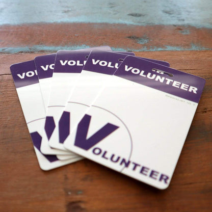 Five "10 Pack - Volunteer Lanyard with Badge Set - Pre-Printed Volunteer Design - Dual Sided" are spread out on a wooden surface. The badges, mostly white with "VOLUNTEER" written prominently in purple letters at the top, are each attached to a lanyard.