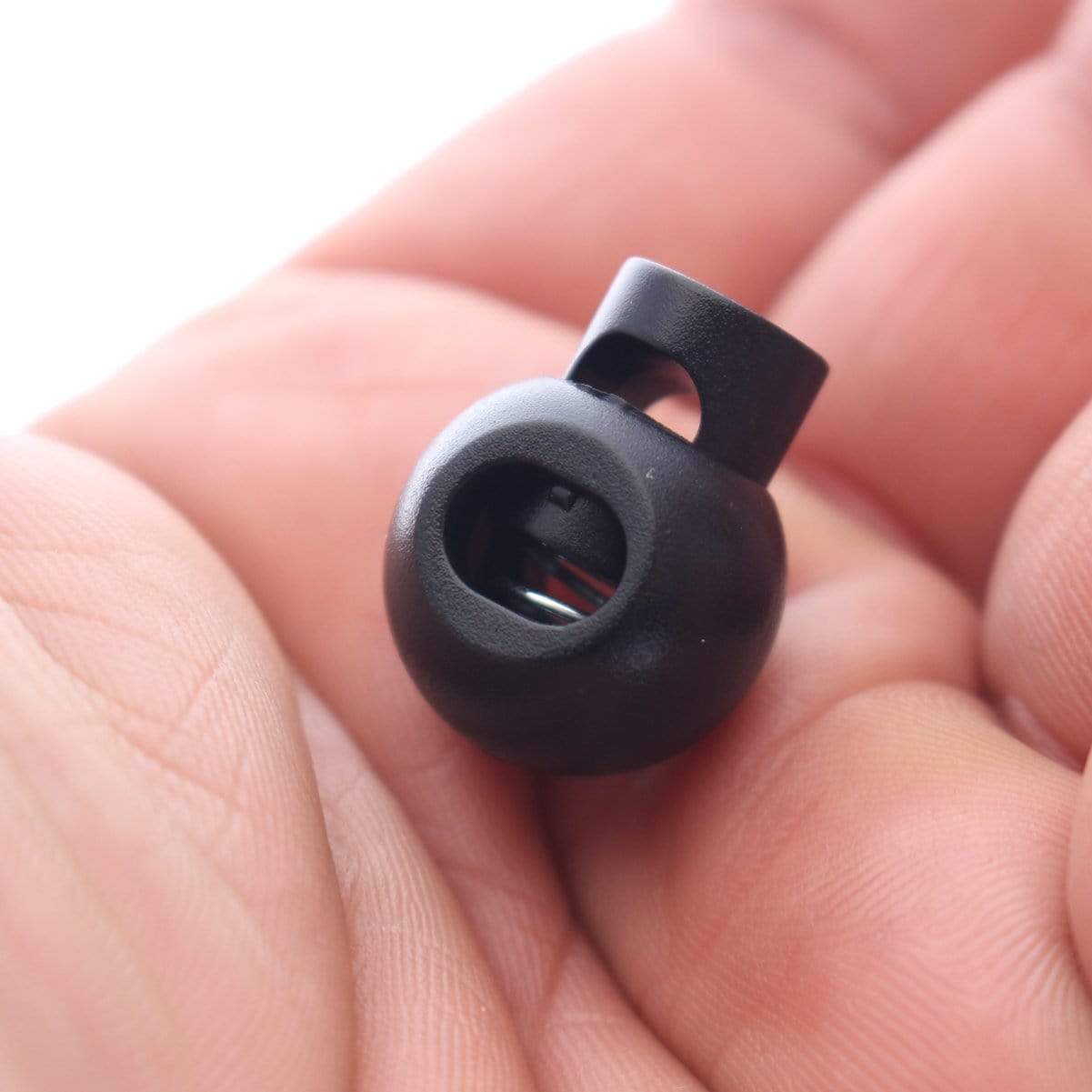 A small, black cylindrical object with an opening and a loop, resembling an Adjustable Cord Lock - Round Ball Style - Single Hole End Toggle for DIY Projects (2135-4001), held in a person's fingertips.