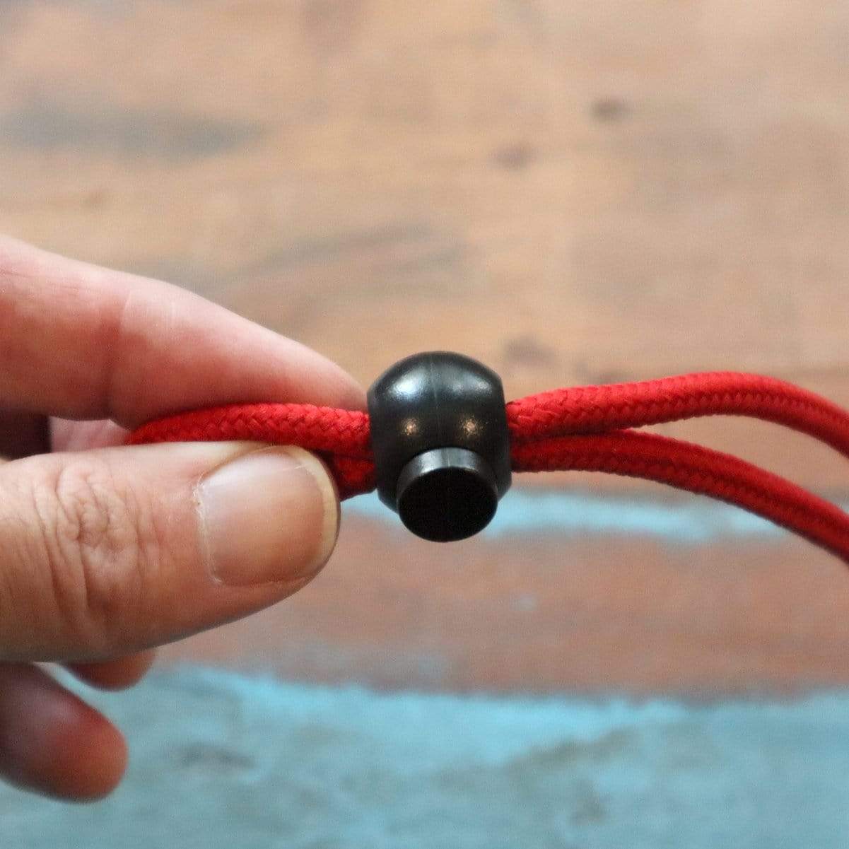 A hand holding an Adjustable Cord Lock - Round Ball Style - Single Hole End Toggle for DIY Projects (2135-4001) threaded through a black toggle fastener against a blurry background.