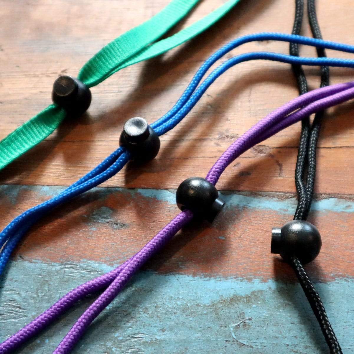 Four Adjustable Cord Lock - Round Ball Style - Single Hole End Toggle for DIY Projects (2135-4001) in green, blue, purple, and black are laid out on a wooden surface.