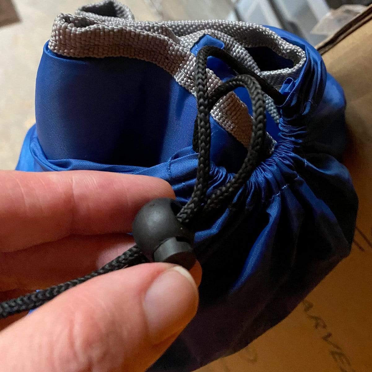 A hand is adjusting an Adjustable Cord Lock - Round Ball Style - Single Hole End Toggle for DIY Projects (2135-4001) on a blue fabric bag with grey accents.