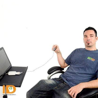 A man sits at a desk using a computer mouse with his right hand. He has short dark hair and is wearing a blue t-shirt and jeans. A Lock-A-Reel Self Locking PIV Card Reel is clipped to his waistband. The desk holds a laptop and a smartphone connected to it.