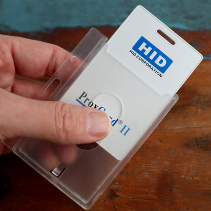 A hand holds a Horizontal 70 - 90 Mil ProxCard II / Thick HID Proximity Card Holder (SPID-PROX-H), showcasing a plastic case containing an HID Corporation ProxCard II identification card on a wooden surface.