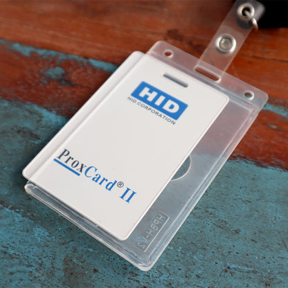 A white HID ProxCard II identification card in a clear plastic Vertical 70 - 90 Mil ProxCard II / Thick HID Proximity Card Holder (SPID-PROX-V) with a black lanyard, placed on a wooden surface.