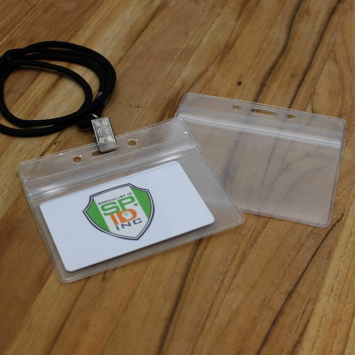 Two clear Horizontal Vinyl Badge Holders with Zipper Top (506-ZHOS-CLR), one empty and the other containing a white card with a green and orange shield logo. Both lie on a wooden surface, with a black lanyard attached to one. The design offers excellent ID card protection.