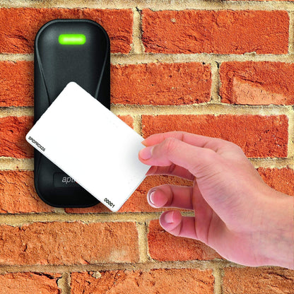 A hand holding an SPIDprox HID Compatible ISO Prox Card near a black card reader with a green light, mounted on a red brick wall. The setup exemplifies modern access control systems utilizing proximity card formats for secure entry.