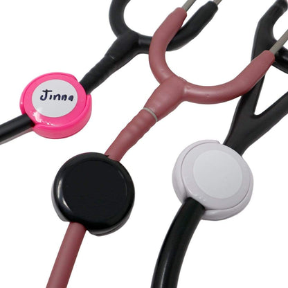Three stethoscopes with black, pink, and white Antimicrobial Stethoscope ID Tags - Hand Write Your Name on Label to Personalize - Adjustable Tube Size to Fit Most Steths. The pink tag has the name "Jinna" written on it, featuring an adjustable size for a perfect fit.