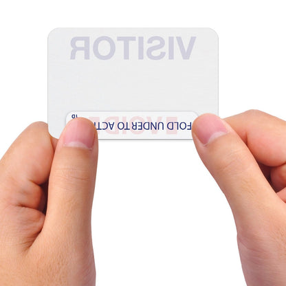 Two hands hold a visitor badge with a peel-off label, revealing text that says "FOLD UNDER TO ACTIVATE." These Visitor Manual ONEstep Quick Tab TimeBadge, Box of 500 (P/N T2023) ensure easy identification and security throughout the premises.