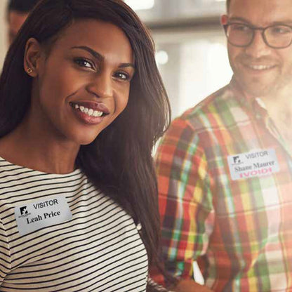 Two people, a woman and a man, wearing Visitor Manual ONEstep Quick Tab TimeBadge, Box of 500 (P/N T2023) are smiling. The woman's badge shows "Leah Price," and the man's badge shows "Shane Warner." They stand closely, facing the camera.