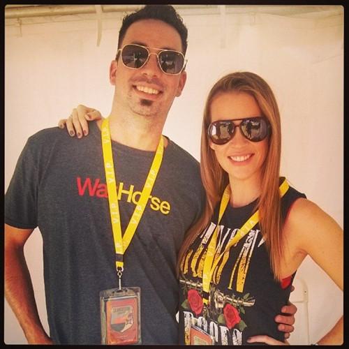 Two people wearing sunglasses and yellow event lanyards with Vertical Oversized 4X6 Vinyl ID Badge Holder (XL46V) smile and pose together against a white background. The man sports a gray "War Horse" shirt, while the woman rocks a "Guns N' Roses" tank top.