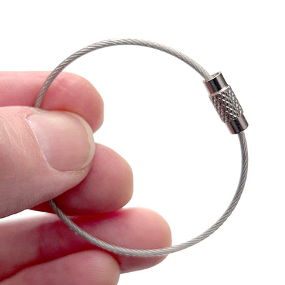 A hand holding a Wire Loop Luggage Tag Holder - 6 inch Metal Stainless Steel Cable Ring Connector for Keys or Luggage Tags with a small metal closure. Sold by Specialist ID.